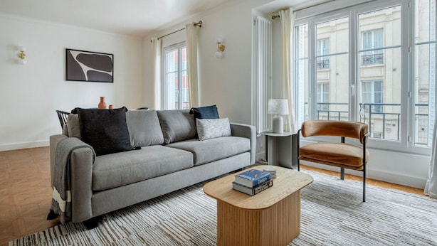 A modern living room with a grey sofa, a wooden coffee table with books, a brown chair, and large windows offering a view of the city. - Blueground | Jules-Joffrin, furnished & equipped (Paris)