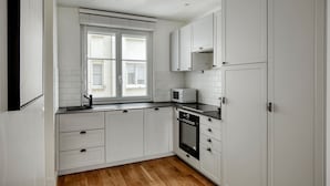 A compact kitchen with white cabinets, a microwave, a stove, and a window with blinds. - Blueground | Jules-Joffrin, furnished & equipped (Paris)