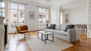 A spacious living room with a sofa, armchair, and coffee table, large windows, and a chandelier. - Blueground | Neuilly, nr dining, gardens & shops (Paris)