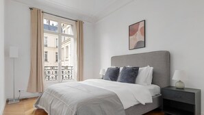 A bedroom with a bed, bedside tables, a window with curtains, and a city view. - Blueground | Neuilly, nr dining, gardens & shops (Paris)