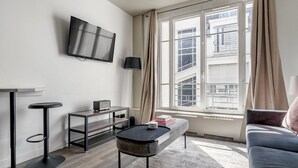 A modern living room with a flat-screen TV, a small table with a speaker, a sofa, and a large window with curtains. - Blueground | Canal Saint Martin, near shops (Paris)