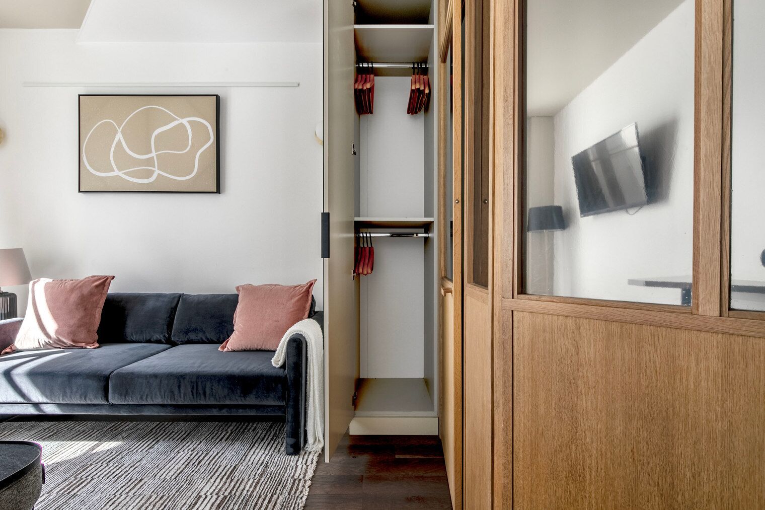 A modern living room with a dark grey sofa, two pink cushions, a wooden cabinet with a TV, and a wall-mounted abstract art piece.
