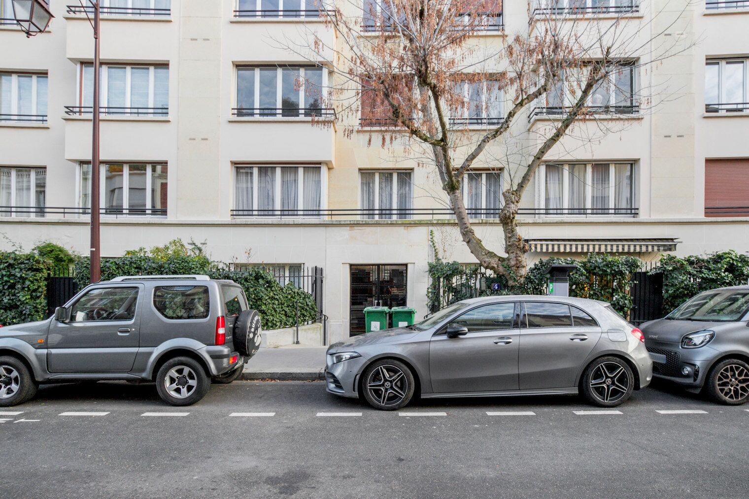 Uma fila de carros estacionados em frente a um prédio residencial com várias andares, sacadas e uma árvore em primeiro plano.
