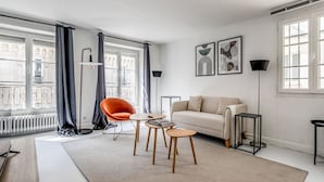 Ein modernes Wohnzimmer mit einer Couch, einem roten Sessel, einem Couchtisch und einer Stehlampe. Es gibt zwei Fenster mit Vorhängen, eine Heizung und Kunstwerke an der Wand.