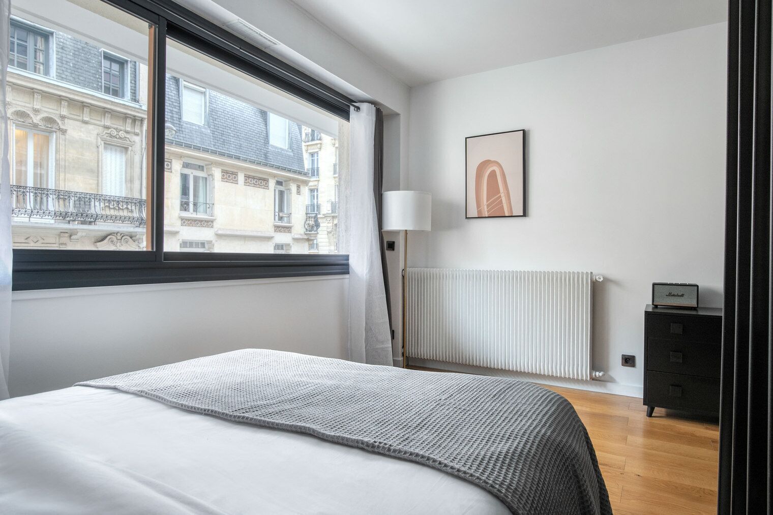 Une chambre à coucher comprenant un lit, un radiateur, une lampe, un tableau encadré et une fenêtre donnant sur des immeubles.