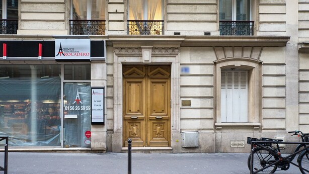 A building facade with a large wooden door, a storefront with a sign for 'Agence Immobilier Roca Déro', and a bicycle parked outside.