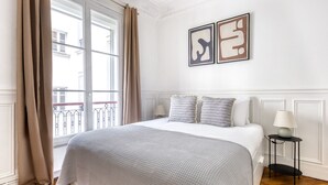 A bedroom with a bed, a window with curtains, a nightstand with a lamp, and two wall art pieces.