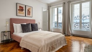 A bedroom with a large bed, a nightstand, a lamp, and two framed artworks on the wall. - Blueground | Saint Georges, nr Palais Garnier (Paris)