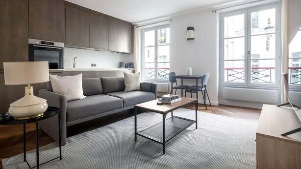 A modern living room with a grey sofa, a coffee table, and a lamp. There is a kitchen area with cabinets and a microwave. The room has large windows with a view of buildings outside. - Blueground | Saint-Honoré, nr opera (Paris)