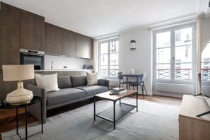 A modern living room with a grey sofa, a coffee table, and a lamp. There is a kitchen area with cabinets and a microwave. The room has large windows with a view of buildings outside. - Blueground | Saint-Honoré, nr opera (Paris)