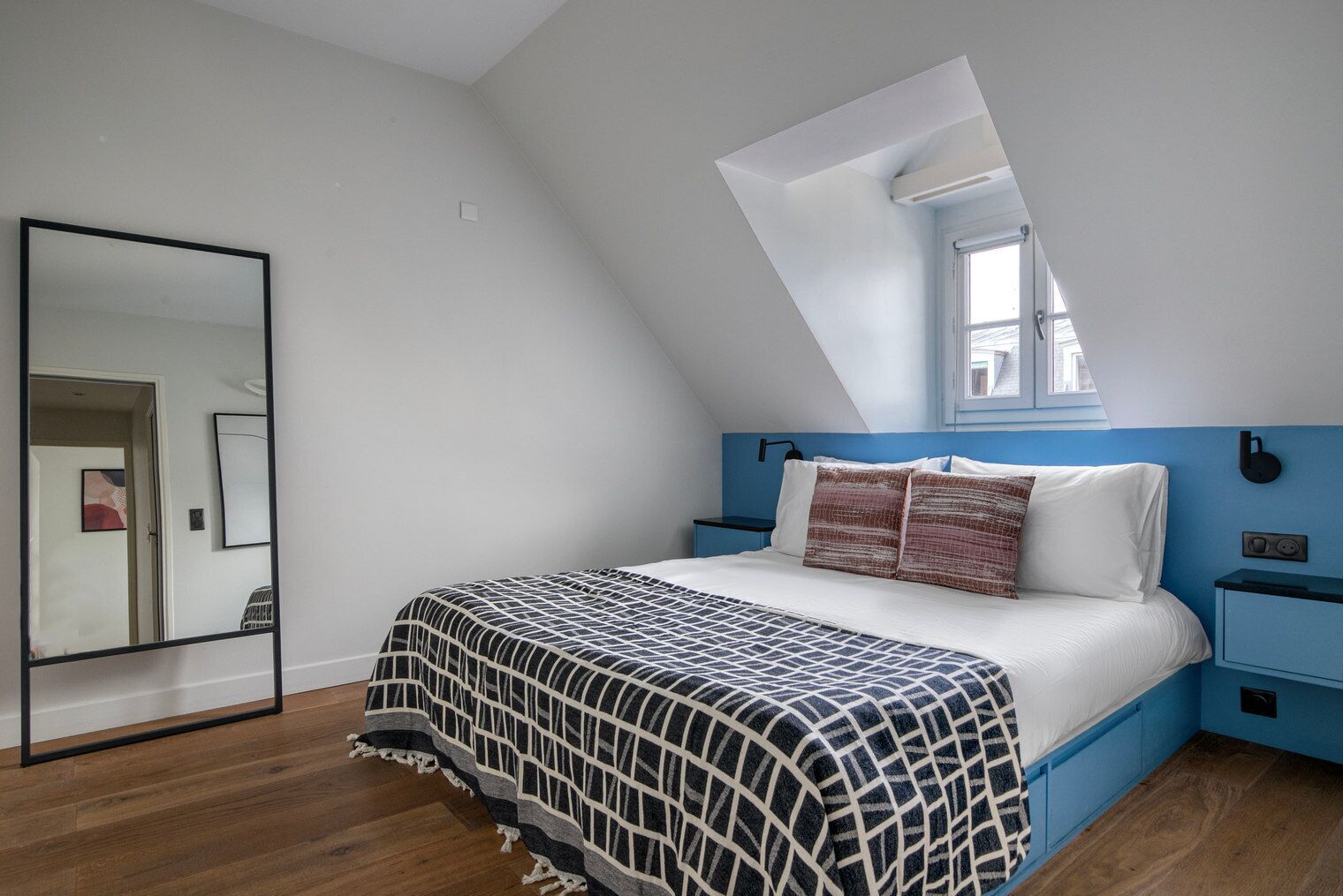 A bedroom with a sloped ceiling, a bed with a patterned blanket, a nightstand, a wall-mounted lamp, and a window with a view of buildings.