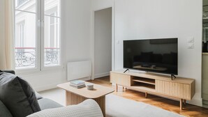 A modern living room with a large flat-screen TV on a wooden stand, a grey sofa, a small coffee table with books and a plate, and a large window with a balcony view. - Blueground | Pigalle, washer, nr festive area (Paris)