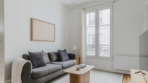 A modern living room with a grey sofa, a wooden coffee table, and a large window with a view of buildings. - Blueground | Pigalle, washer, nr festive area (Paris)