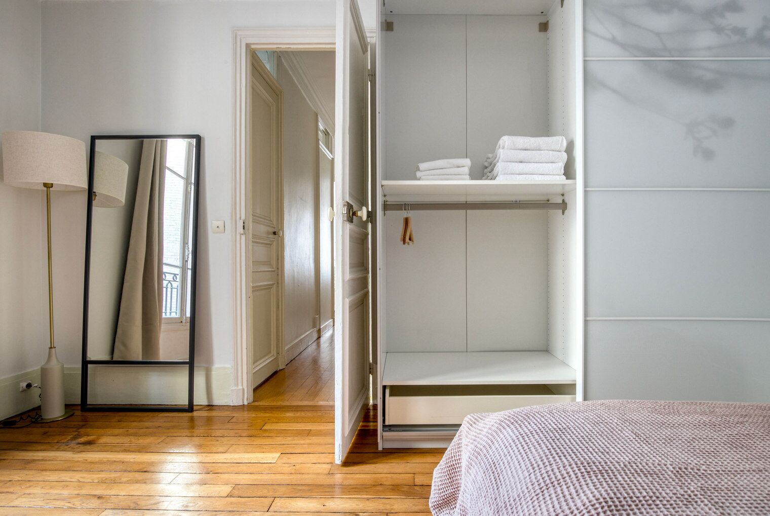 A bedroom with a wooden floor, a bed with a pink blanket, a wardrobe with hanging clothes, and a mirror.