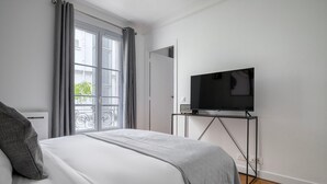 A bedroom with a bed, a TV on a stand, a window with curtains, and a view of a balcony. - Blueground | Victor Hugo, nr Trocadero Garden (Paris)