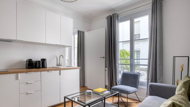 A modern kitchen with white cabinets, a wooden countertop, and a coffee maker. There is a glass table with a book, a grey armchair, and a grey sofa. A window with grey curtains is visible. - Blueground | Victor Hugo, nr Trocadero Garden (Paris)
