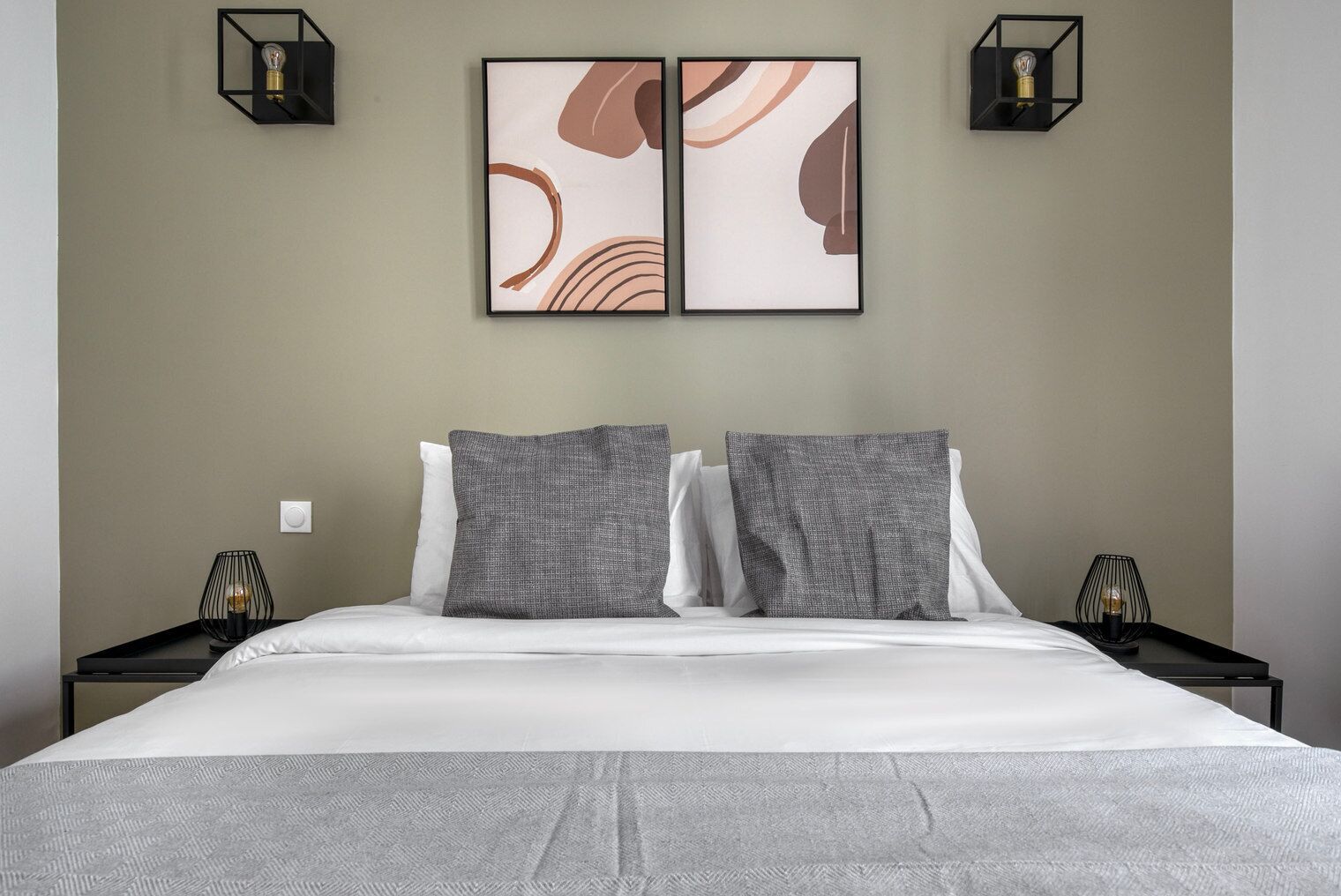 Une chambre d’hôtel comprenant un lit, deux tables de chevet, un luminaire fixé au mur et une œuvre d’art abstraite encadrée.