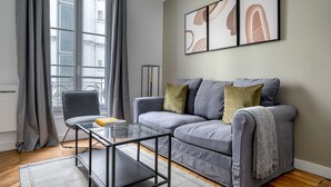 A modern living room with a grey sofa, a glass coffee table, and a grey armchair. There are three cushions on the sofa, a grey throw blanket, and a floor lamp. The room has a large window with grey curtains. - Blueground | Victor Hugo, nr Trocadero Garden (Paris)