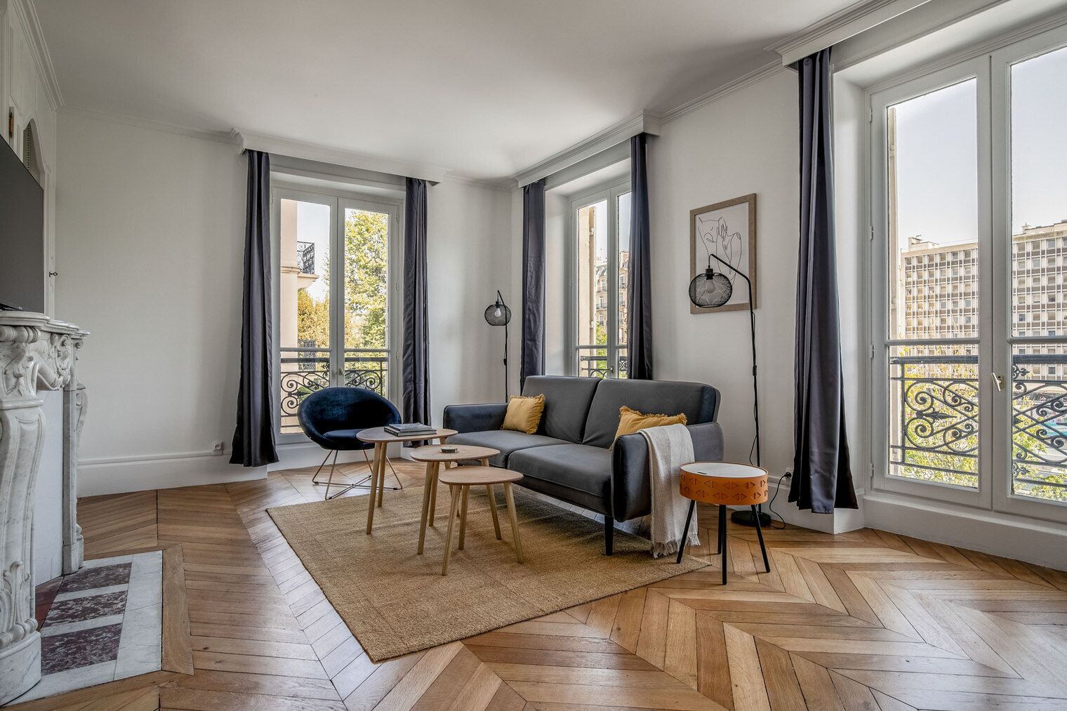 Una sala de estar moderna con un sofá gris, un sillón azul y una mesita. Se observan ventanas amplias con cortinas, una lámpara de pie y un adorno en la pared.