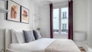 A neatly arranged bedroom with a bed, pillows, a bedside table, and a window with curtains. - Blueground | Canal St Martin, near cafes & shops (Paris)