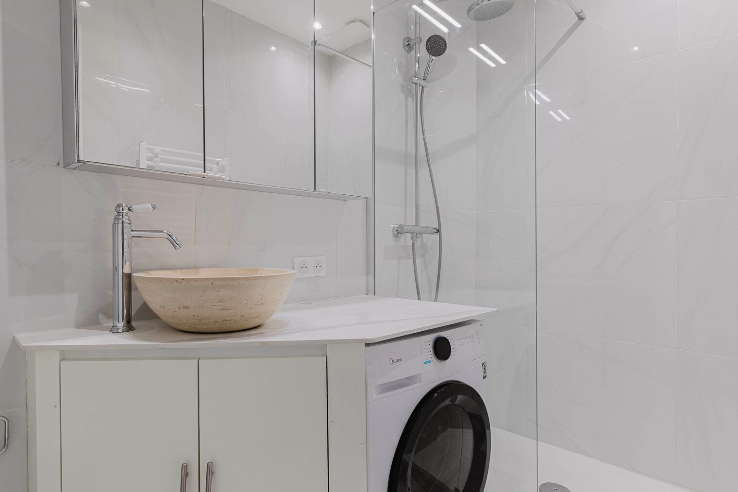 A modern bathroom with a glass shower enclosure, a beige ceramic sink, and a front-loading washing machine.