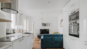 A modern kitchen with white cabinetry, a built-in oven, and a dining area with a blue sofa. - Blueground | Commerce, close to offices & shops (Paris)