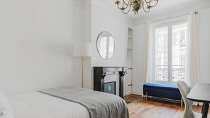 A bedroom with a bed, a desk, a chair, a fireplace, and a window with a view of buildings. - Blueground | Commerce, close to offices & shops (Paris)