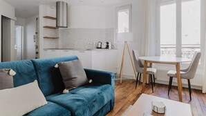 A modern living room with a blue sofa, a dining table with chairs, a kitchen area with white cabinets, and a large window with a view. - Blueground | Commerce, close to offices & shops (Paris)