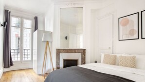 A bedroom with a large bed, a fireplace, a floor lamp, and a view through a window.