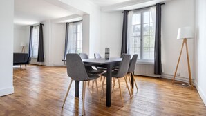 Ein geräumiges Zimmer mit Holzboden, einem runden Tisch mit Stühlen und großen Fenstern mit Vorhängen.