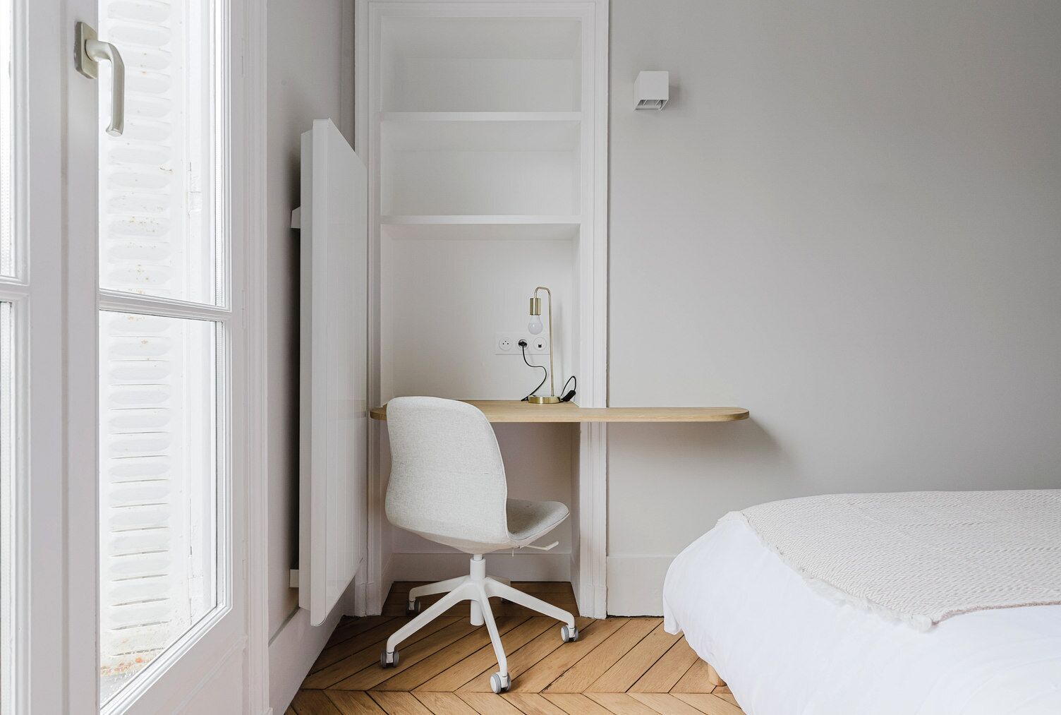 Ein Schlafzimmer mit Bett, Schreibtisch mit Stuhl, einem Fenster mit Jalousien und einer Wandleuchte.