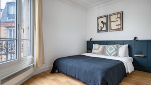 Une chambre avec une grande fenêtre, un lit doté d’une tête de lit sombre et deux œuvres d’art encadrées accrochées au mur.
