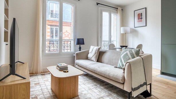 Un salon moderne avec un canapé, une table basse et un téléviseur. De grandes fenêtres sont présentes, ainsi que des rideaux, une lampe et un tableau encadré accroché au mur.