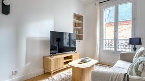 Un salon moderne avec un téléviseur à écran plat posé sur un meuble en bois, un canapé beige, une petite table basse et une fenêtre donnant sur un bâtiment en briques.