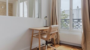A small, bright room with a wooden desk and a chair, a large window with curtains, and a view of a balcony with a metal railing.