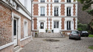 A two-story building with a brick facade and white trim. There are steps leading to a door with a small garden area in front. Two cars are parked in the driveway.