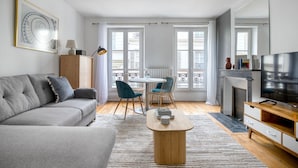 Ein modernes Wohnzimmer mit einem grauen Sofa, einem Holztisch, einer Essecke mit einem weißen Tisch und blauen Stühlen sowie einem Fernseher auf einem Holzschrank.