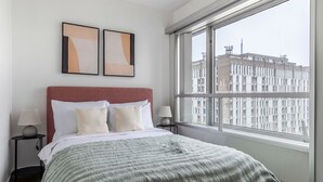 A bedroom with a large window, a bed with white and beige bedding, two bedside tables with lamps, and two abstract paintings on the wall. - Blueground | La Motte-Picquet Grenelle, nr seine (Paris)