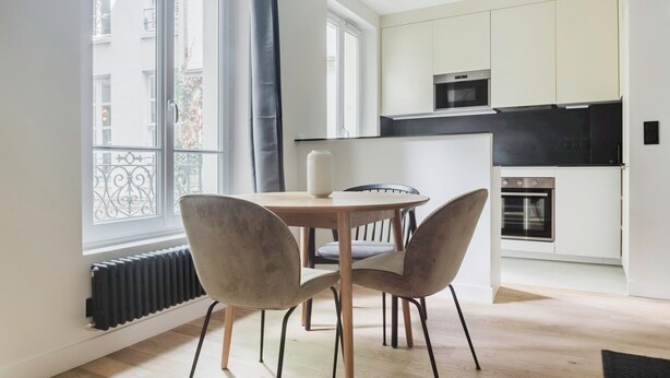 A modern kitchen with white cabinets, a black countertop, and stainless steel appliances. There is a dining area with a wooden table and two chairs. - Blueground | Jardin des Plantes, furnished (Paris)