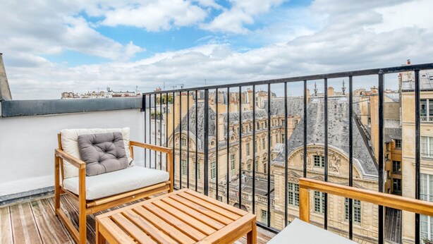 Un balcon avec une chaise et une table en bois, offrant une vue sur des bâtiments historiques et un ciel dégagé.