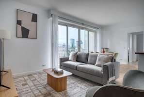A modern living room with a grey sofa, a wooden coffee table, and a view of the city through large windows. - Blueground | Pont de Neuilly, balcony, nr shops (Paris)