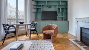 Een woonkamer met een open haard, een televisie bevestigd aan een blauwgroene muur, en een eethoek met een tafel en stoelen.