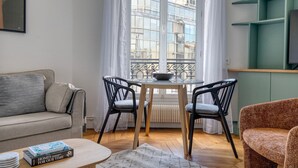 Een gezellige woonkamer met een bank, een klein tafeltje, twee stoelen, een radiator en een raam met uitzicht op gebouwen.