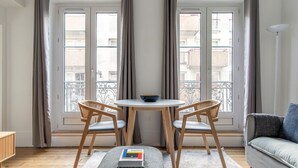 A modern living room with a round table, two chairs, a sofa, and a view of the city through large windows. - Blueground | Buttes-Chaumont, walk to garden (Paris)