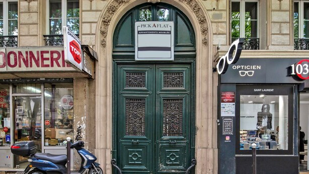 A building with a green ornate door labeled "PICK A FLAT" and a storefront with a sign reading "OPTICIEN 103". - Blueground | Buttes-Chaumont, walk to garden (Paris)