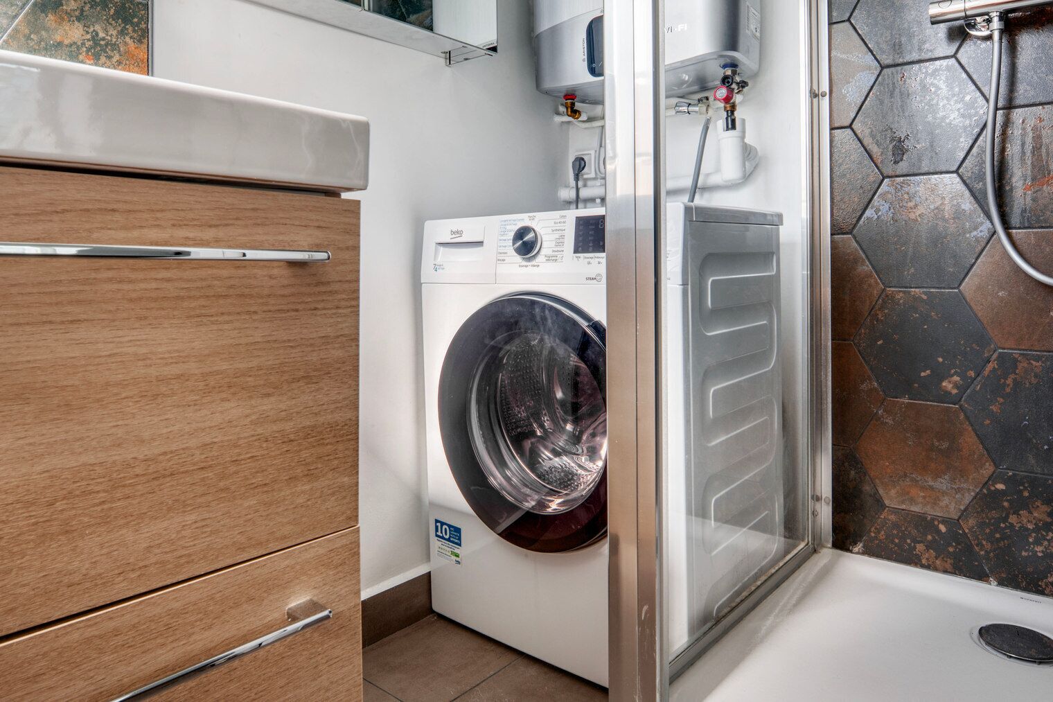 A compact laundry room with a front-loading washing machine, a wooden cabinet, and a shower area with hexagonal tiles.