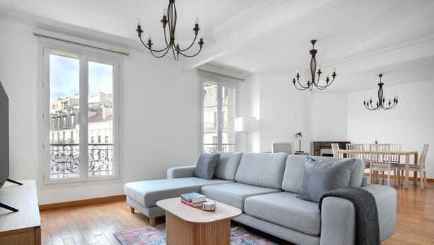 A modern living room with a grey sectional sofa, a wooden coffee table, and a patterned rug. There are two chandeliers, a dining area with a wooden table and chairs, and large windows with a view of buildings. - Blueground | La Motte-Picquet Grenelle, near shops (Paris)