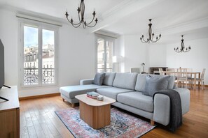 A modern living room with a grey sectional sofa, a wooden coffee table, and a patterned rug. There are two chandeliers, a dining area with a wooden table and chairs, and large windows with a view of buildings. - Blueground | La Motte-Picquet Grenelle, near shops (Paris)