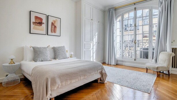 Une chambre avec un grand lit, une fenêtre avec des rideaux, une chaise et une petite table.