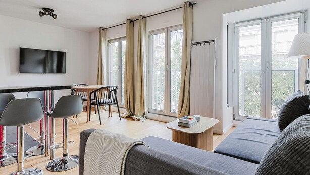 En moderne stue med sofa, spiseområde med stole, et fjernsyn ophængt på væggen og store vinduer med gardiner.